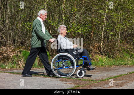 Retired husband taking disabled elderly wife in wheelchair for a walk in the park on a sunny day in spring Stock Photo