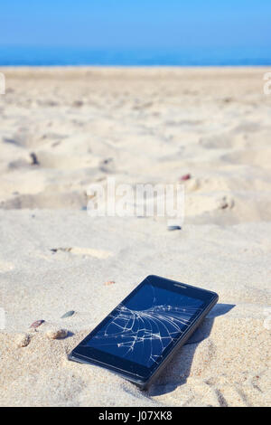 Close up picture of a mobile phone with broken screen in sand on a beach, selective focus. Stock Photo