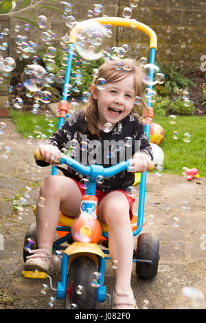 Four-year-old girl on toy bicycle in garden with bubbles blowing around. England. Stock Photo