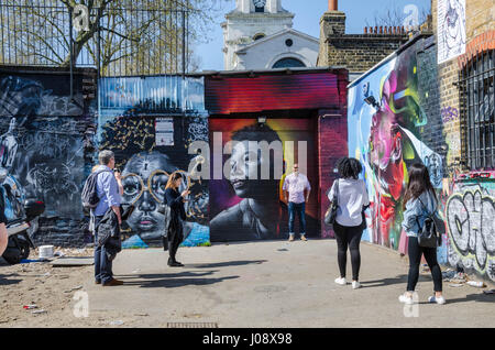 Tourists admire and pose for photographs with the vibrant, colourful street are which covers the walls of a parking area off Brick Lane in London. Stock Photo
