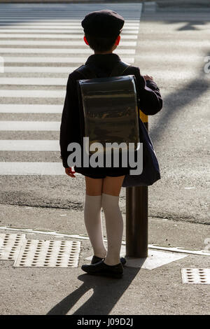 A Japanese school boy wearing an old fashioned school uniform and carrying a satchel waits to cross the road in Yokohama, Kanagawa, Japan Stock Photo