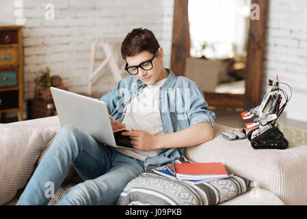 Keep silent please. Smiling boy holding laptop on the knees looking at copybook while making some notes Stock Photo