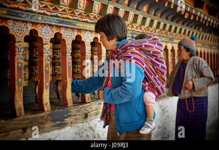 Two women, one carrying a baby, spin prayer wheels on the wall of the Changangkha Lhakhang Temple in Thimphu Bhutan Stock Photo