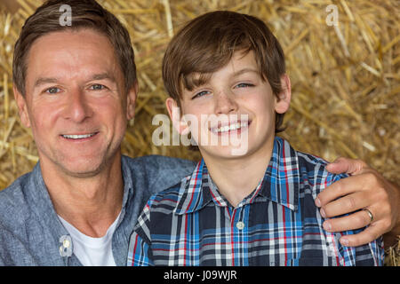 Portrait shot of an attractive, successful and happy middle aged man male sitting on hay bales with his male child boy son in a barn or stables Stock Photo