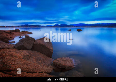 Cloudy blue sunrise over still waters of Jindabyne lake in Snowy mountains of NSW, Australia. The lake is surrounded by snow head mountain ranges. Stock Photo