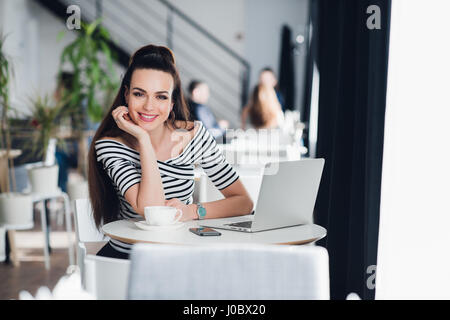 Picture of happy smiling woman using laptop in a cafe and looking at the camera. Stock Photo