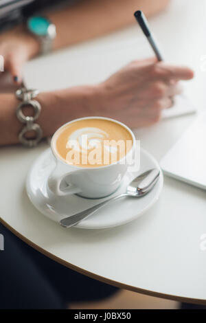 Hands writing on a paper in a notebook. Cup of cappuccino. Laptop nearby. Stock Photo