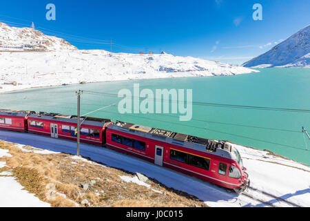 Bernina Express train in the snowy valley surrounded by Lake Bianco, Bernina Pass, Canton of Graubunden, Engadine, Switzerland Stock Photo