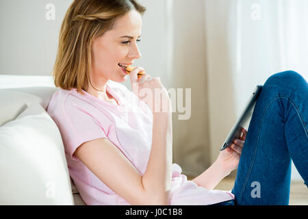 Smiling young woman eating cookie and using digital tablet at home Stock Photo