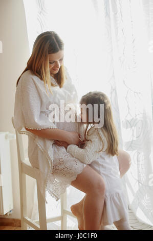 Charming little girl hugging her pregnant mother. The woman in a beautiful white dress sits on a chair, and her little daughter kisses a mother's stom Stock Photo