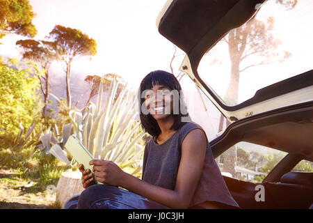 Portrait of young woman sitting in open car boot, holding digital tablet Stock Photo