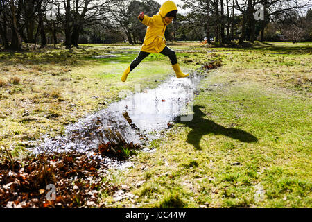 Boy in yellow anorak jumping over puddle in park Stock Photo
