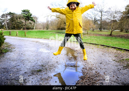 Boy in yellow anorak jumping above puddle in park