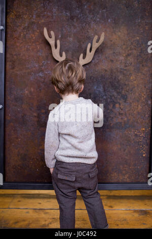 Young boy standing facing wall, cardboard reindeer cut out on wall behind him Stock Photo