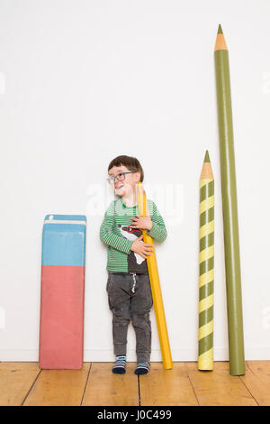 Young boy standing, holding giant size pencil, giant stationery leaning on wall beside him Stock Photo