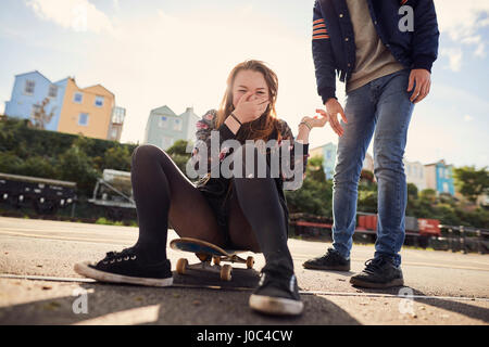 Two friends fooling around outdoors, young woman sitting on skateboard, laughing, low section, Bristol, UK Stock Photo