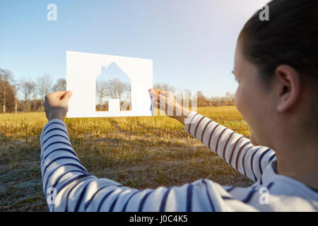 Woman standing in rural setting, holding out paper with cut-out of house Stock Photo