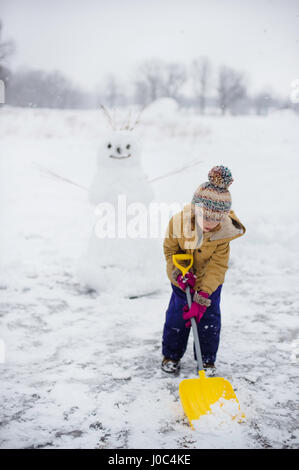 Girl shovelling snow in front of snowman, Lakefield, Ontario, Canada Stock Photo