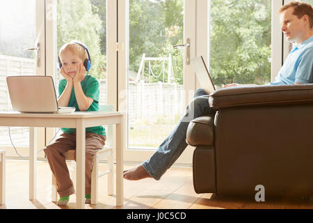 Boy at desk and father on sofa using laptops Stock Photo