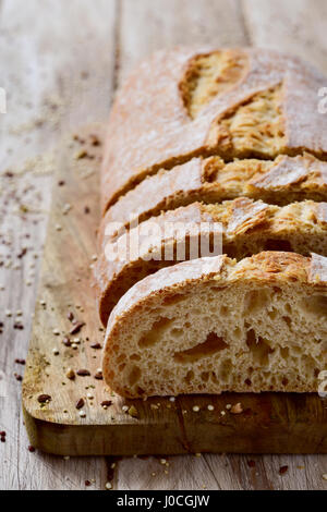 closeup of a loaf of spelt bread cut in some slices on a chopping board, placed on a white rustic wooden table Stock Photo