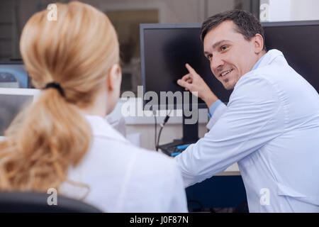 With colleagues. Pleasant happy nice doctor smiling and pointing at the monitor while working together with his colleague Stock Photo