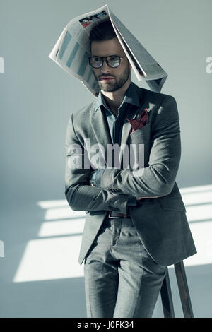 Handsome stylish man in eyeglasses sitting on stool with newspaper on head Stock Photo