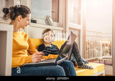 Mother and daughter sitting on yellow sofa and talking while using laptop and tablet Stock Photo
