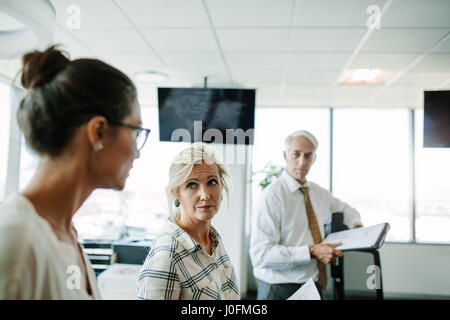Mature businesswoman listening t her colleague with man standing in background. Business people having a informal meeting in modern office. Stock Photo