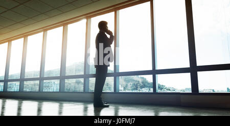 Full length of entrepreneur standing by window and talking on mobile phone. Man looking outside the office window and making a phone call. Stock Photo