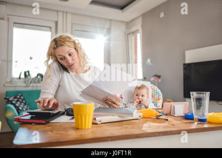 Mother holding letter, using calculator, talking on smart phone at home office and taking care of her baby Stock Photo