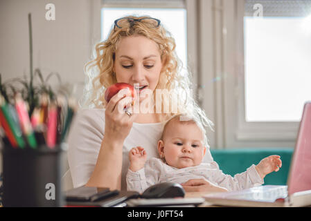 Mother eating red apple and using laptop at home office with baby in her arms Stock Photo