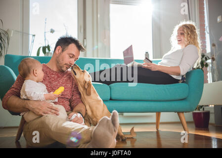 Father sitting on the floor and with baby in his arms playing with dog while wife sitting on sofa and doing online shopping with credit card Stock Photo