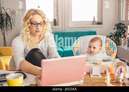 Mother wearing eyeglasses working at home office on laptop and taking care of her baby Stock Photo