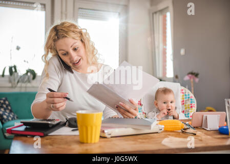 Mother holding letter, writing notes, talking on smart phone at home office and taking care of her baby Stock Photo