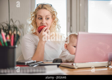 Mother eating red apple and using laptop at home office with baby in her arms Stock Photo