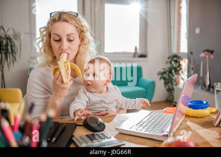 Mother eating bananas and holding baby in her arms at home office Stock Photo