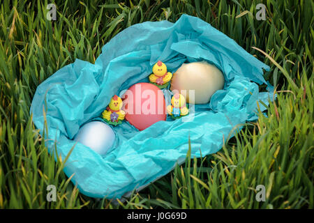 Three toy chickens and three easter eggs on the grass in a blue basket. Composition. Stock Photo