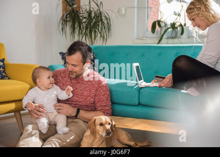 Father sitting on the floor and holding baby in his arms while wife sitting on sofa and doing online shopping with credit card Stock Photo