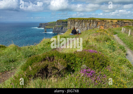 A couple sat on the cliff edge of the Cliffs of Moher, looking north east towards O'Brien's Tower, County Clare, Ireland Stock Photo