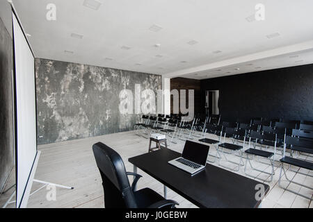 Room for lecture with a lot of dark chairs. Walls are white, loft interior. On the right there is a door. On the background there is a table with a laptop. Stock Photo
