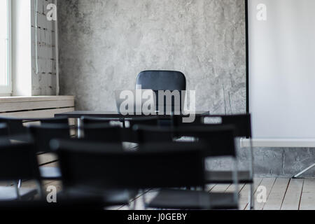 Room for lecture with a lot of dark chairs. Walls are white, loft interior. On the right there is a door. On the background there is a table with a laptop. Stock Photo