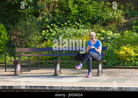 Young woman using mobile phone sitting on bench waiting for train at railway station at Bradford on Avon, Wiltshire in April Stock Photo