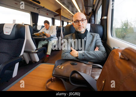 A middle aged male figure sitting on the train wearing jacket and glasses in first class carriage Stock Photo