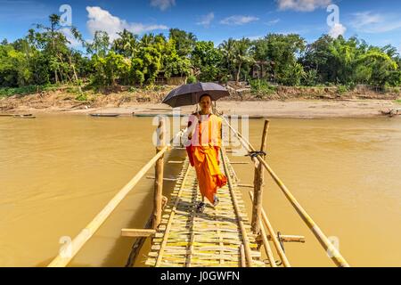 A young Buddhist monk walks across a bamboo bridge over Nam Khan in Luang Prabang Laos