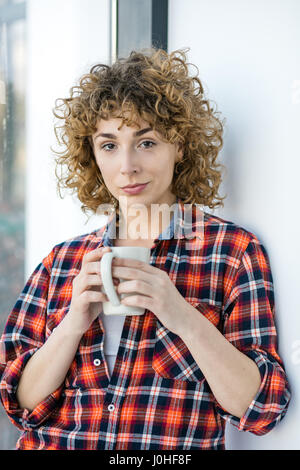 young natural curly woman casual dressed in a checkered shirt lying to a window inside his home and holding a cup of coffee or tea in hands Stock Photo