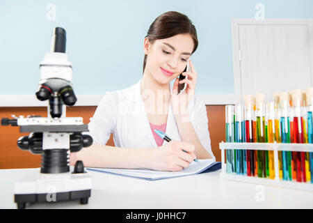 Young female scientist in lab coat talking on smartphone and taking notes in laboratory Stock Photo