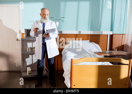 Concentrated senior bearded male doctor making notes in clipboard Stock Photo