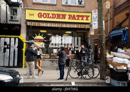 Canada, Toronto. Kensington market, multicultural neighbourhood . Customers eat patties in front of  The Golden Patty bakery. Stock Photo