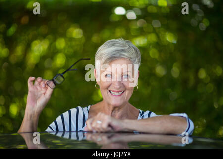 Portrait of senior woman holding eyeglasses while leaning on car roof Stock Photo