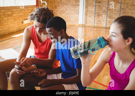 High school girl drinking water while boys using mobile phone in basketball court Stock Photo